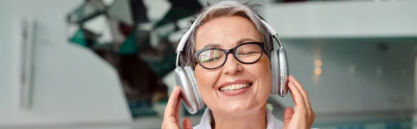 Heureuse femme d'âge moyen dans les lunettes écouter de la musique dans les écouteurs sans fil dans le centre de spa, bannière — Photo de stock