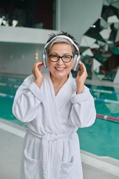 Glückliche Frau mittleren Alters mit Brille, die Musik in drahtlosen Kopfhörern im Wellnesszentrum hört, Rückzug — Stockfoto