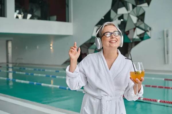 Zufriedene Frau mittleren Alters hört Musik in drahtlosen Kopfhörern und hält Cocktail im Wellness-Center — Stockfoto