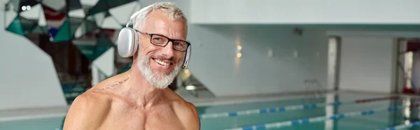 Pleased and tattooed mature man listening music in wireless headphones in spa center, banner — Stock Photo