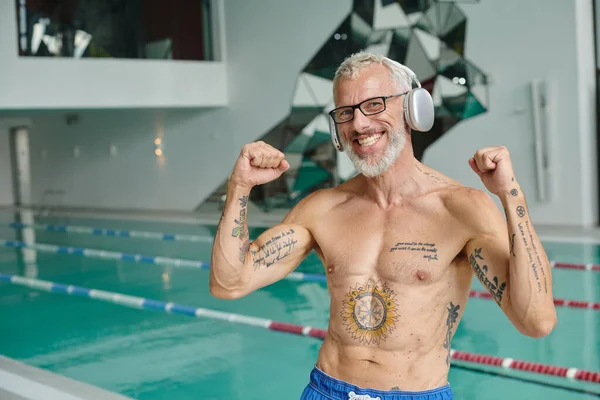 Excité et tatoué homme d'âge moyen écouter de la musique dans les écouteurs sans fil dans le centre de spa, retraite — Photo de stock