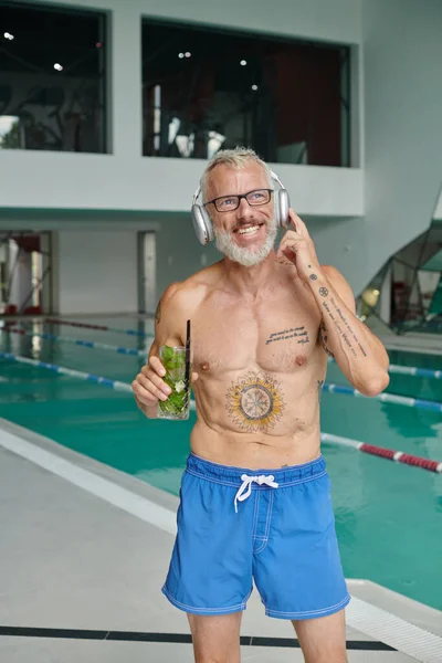 Ritiro, tatuato uomo di mezza età ascoltando musica in cuffia e tenendo cocktail nel centro benessere — Foto stock
