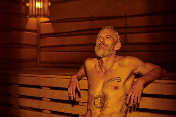 Nachdenklicher und hemdsloser Mann mittleren Alters mit Tätowierungen sitzt in der Sauna, Wellness-Rückzugskonzept — Stockfoto