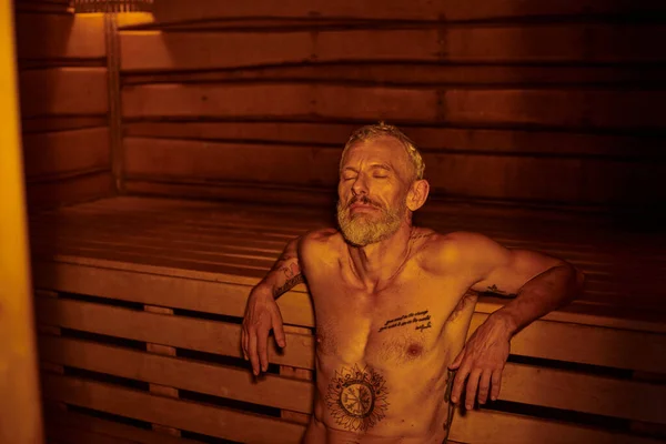 Tranquil, relaxed and shirtless middle aged man with tattoos sitting in sauna, wellness retreat — Stock Photo