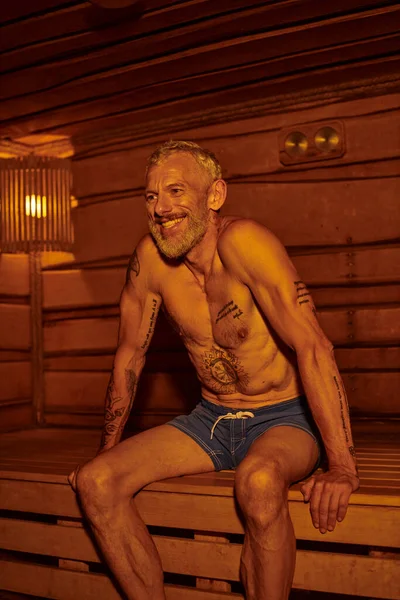 Положительный и без рубашки мужчина среднего возраста с татуировками сидя в сауне, оздоровительный ретрит концепции — стоковое фото