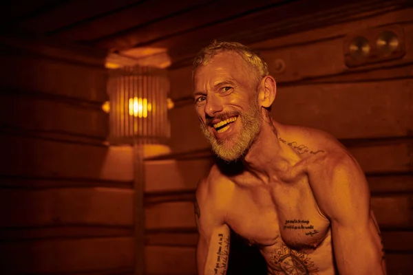 Hombre de mediana edad alegre y sin camisa con tatuajes sentado en la sauna, concepto de retiro de bienestar - foto de stock