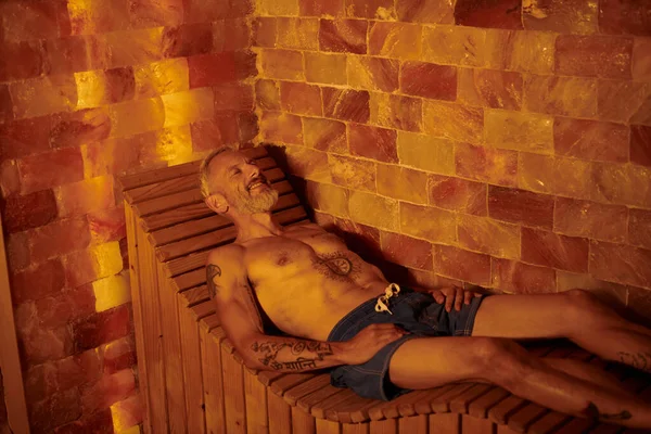Радісний і без сорочки чоловік середнього віку з татуюваннями, що лежать у сауні, концепція оздоровчого відступу — стокове фото