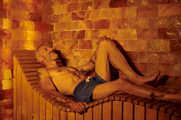 Розслаблений і без сорочки чоловік середнього віку з татуюваннями, що лежать у сауні, концепція оздоровчого відступу — стокове фото