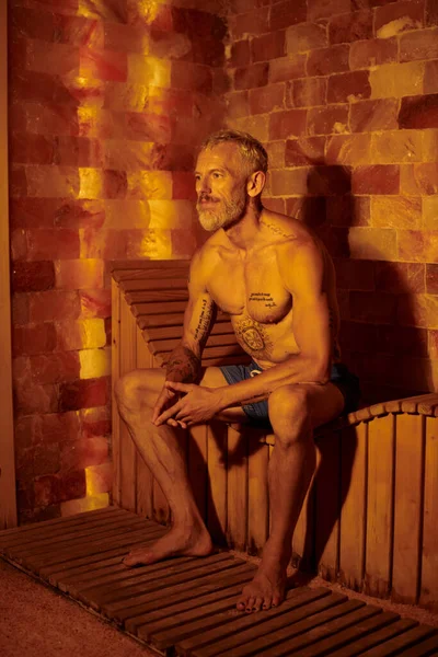 Расслабленный и без рубашки мужчина среднего возраста с татуировками, сидящий на скамейке в сауне, оздоровительная концепция — стоковое фото