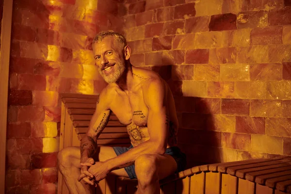 Щасливий і без сорочки чоловік середнього віку з татуюваннями, сидячи на лавці в сауні, концепція оздоровлення — стокове фото
