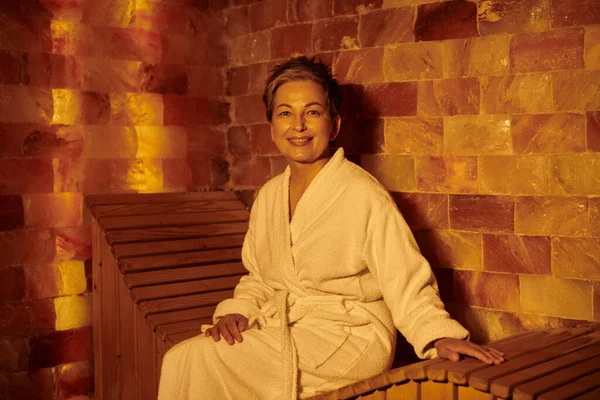 Feliz mujer de mediana edad en bata blanca sentado en el banco en la sauna, spa concepto de bienestar, retiro - foto de stock