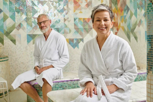 Happy middle aged couple in white robes sitting together in marble sauna, spa wellness concept — Stock Photo