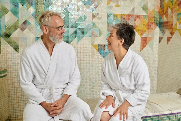 Happy middle aged couple in white robes chatting and sitting in marble sauna, spa wellness concept — Stock Photo