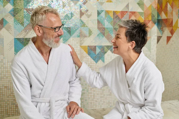 Happy middle aged couple in white robes chatting and sitting in sauna, spa wellness concept, hammam — Stock Photo
