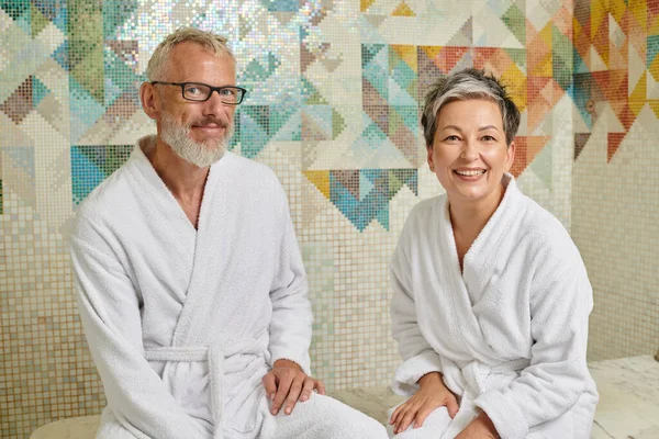 Cheerful middle aged couple in white robes sitting in sauna, spa and wellness concept, hammam — Stock Photo