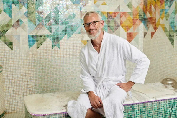 Cheerful middle aged man in white robe sitting in sauna, spa and wellness concept, marble hammam — Stock Photo
