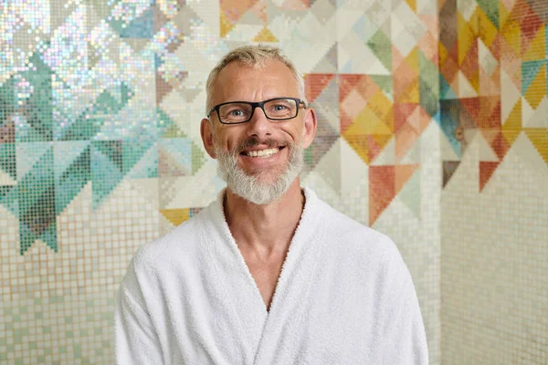Joyful middle aged man in white robe sitting in sauna, spa and wellness concept, marble hammam — Stock Photo