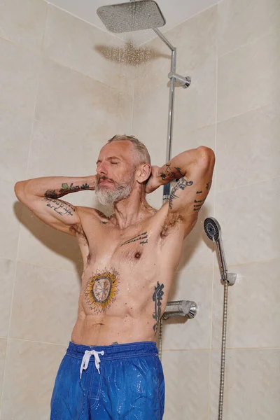 Hombre de mediana edad sin camisa con tatuajes y ojos cerrados tomando ducha, higiene personal - foto de stock