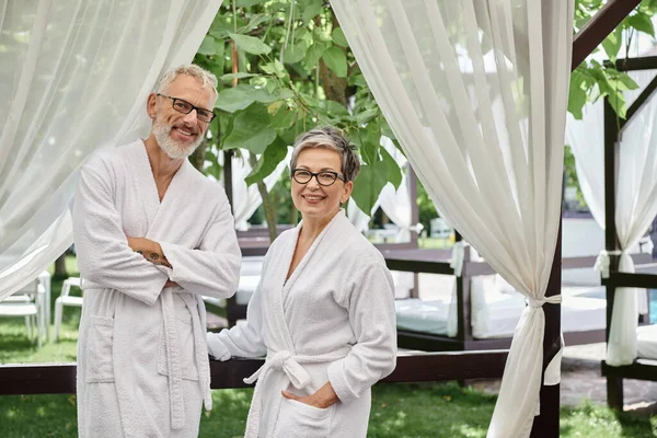 Happy middle aged couple in glasses and robes looking at camera, posing, wellness retreat concept — Stock Photo