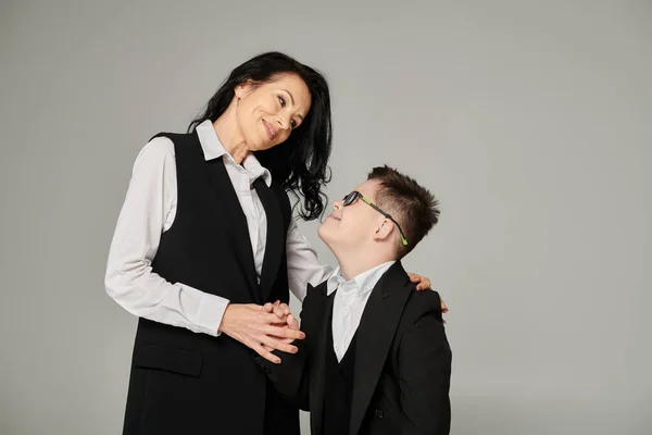 Schoolboy with down syndrome and middle aged mother in formal wear smiling at each other on grey — Stock Photo