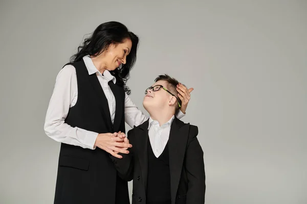Mulher em traje de negócios e filho com síndrome de down em uniforme escolar olhando um para o outro em cinza — Fotografia de Stock