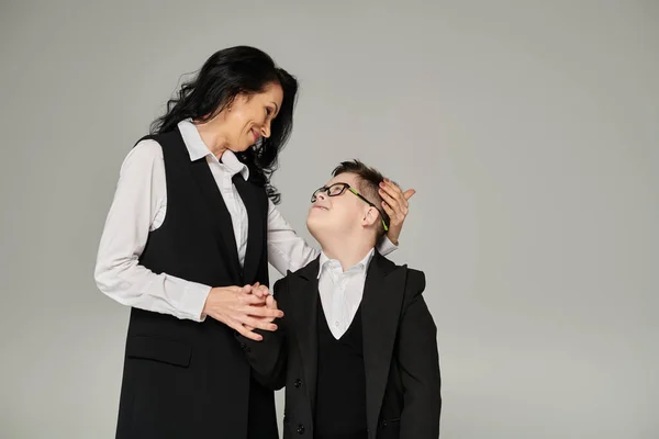 Woman in business attire and son with down syndrome in school uniform looking at each other on grey — Stock Photo