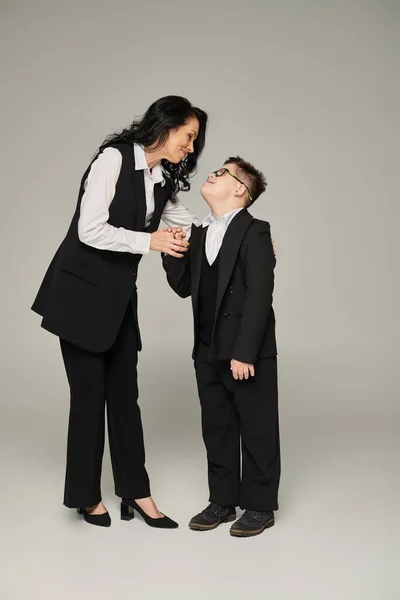 Mujer de negocios e hijo con síndrome de Down en uniforme escolar tomados de la mano y sonriendo en gris - foto de stock