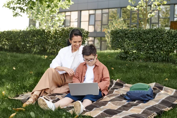 Boy with down syndrome, in eyeglasses, sitting with laptop in park near happy mother, e-learning — Stock Photo