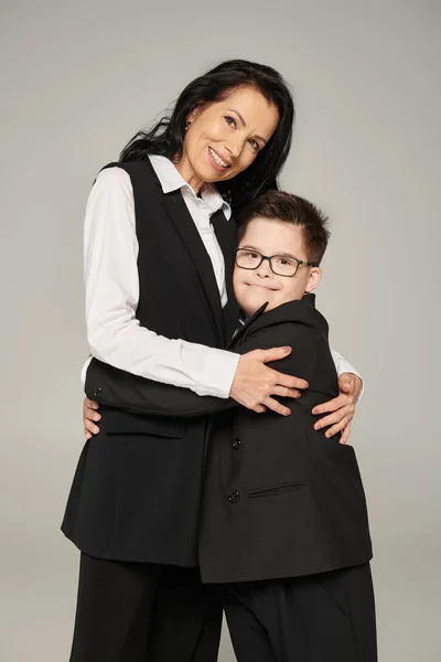 Cheerful schoolboy with down syndrome embracing mother in formal wear and looking at camera on grey — Stock Photo