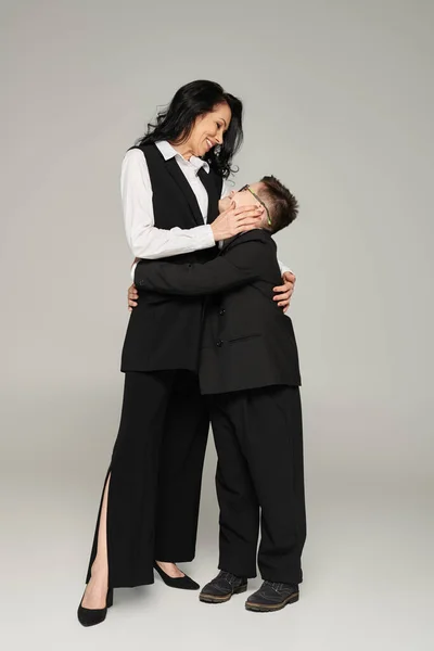Boy with down syndrome, in school uniform, and woman in formal wear embracing on grey, unique family — Stock Photo