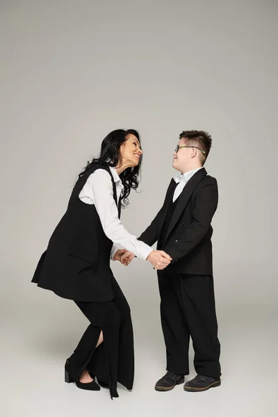 Freudige Geschäftsfrau hält Händchen ihres Sohnes mit Down-Syndrom in Schuluniform auf grau, in voller Länge — Stockfoto