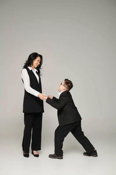 Schoolboy with mental disability holding hands of happy mother in formal wear on grey, full length — Stock Photo