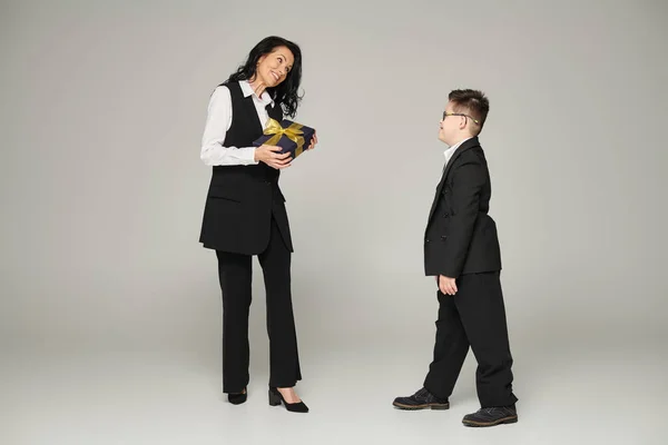 Женщина в деловой одежде держит подарочную коробку рядом с сыном с синдромом Дауна в школьной форме на сером — стоковое фото