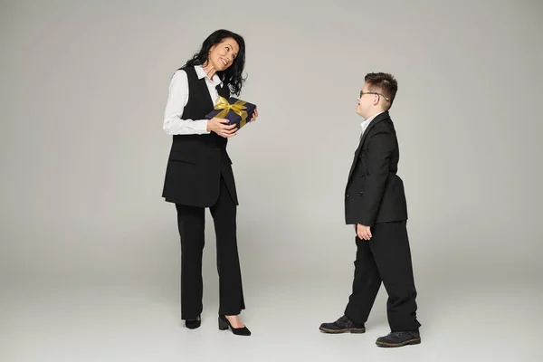 Woman in business attire holding gift box near son with down syndrome in school uniform on grey — Stock Photo