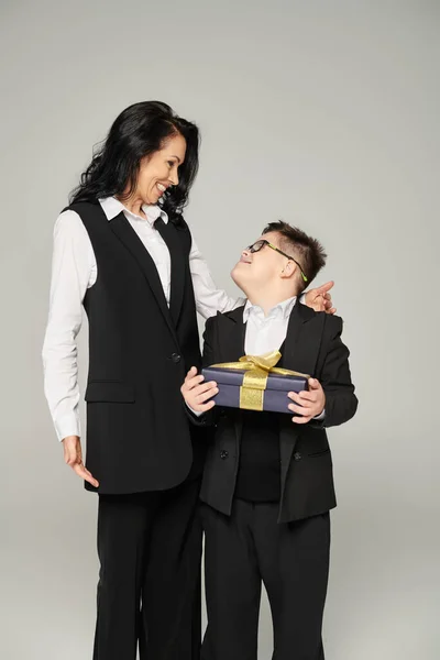 Pleased schoolboy with down syndrome holding present near mother in formal wear on grey, happiness — Stock Photo