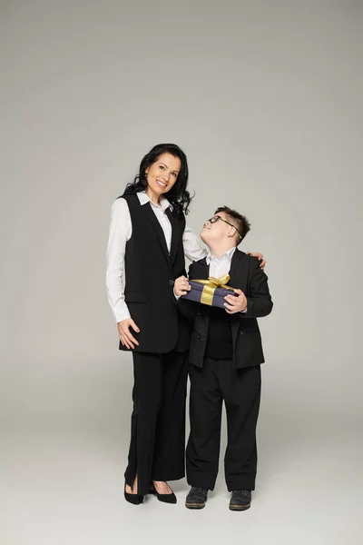 Mulher em traje de negócios sorrindo para a câmera perto do filho com síndrome de down segurando caixa de presente no cinza — Fotografia de Stock