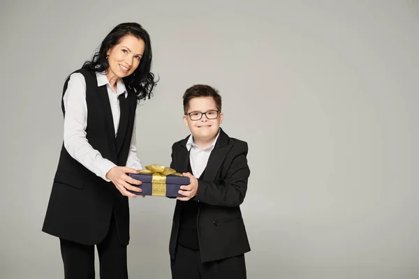 Alegre madre e hijo con síndrome de Down en uniforme escolar sosteniendo caja de regalo y sonriendo en gris - foto de stock