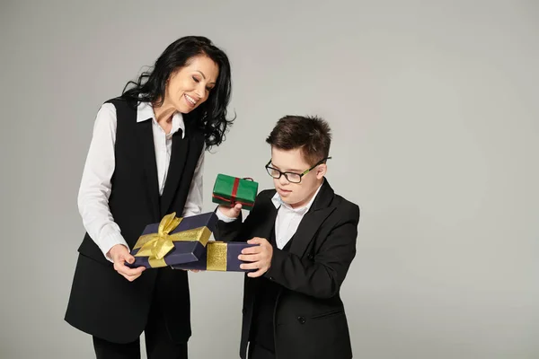 Preteen schoolboy with down syndrome opening gift box near happy mother in formal wear on grey — Stock Photo