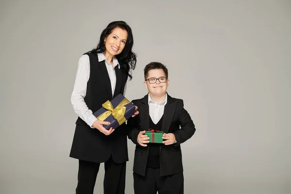 Woman in business attire and boy with down syndrome in school uniform holding gift boxes on grey — Stock Photo