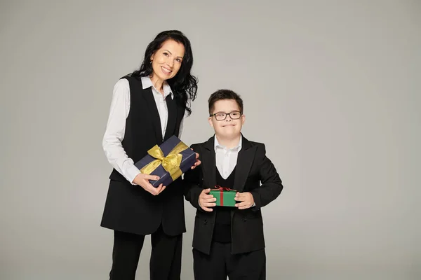 Donna in abito da lavoro e ragazzo con la sindrome di Down in uniforme scolastica contenente scatole regalo su grigio — Foto stock