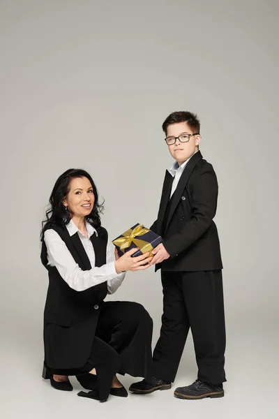 Freudige, elegante Mutter und ihr Sohn mit Down-Syndrom halten eine Geschenkschachtel in der Hand und blicken in die Kamera auf grau — Stockfoto