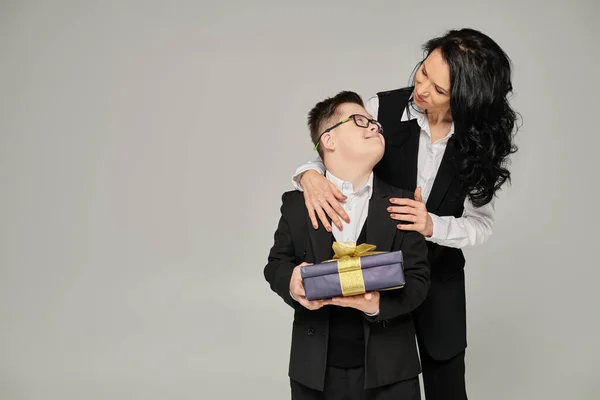 Счастливая женщина в формальной одежде обнимает сына с синдромом Дауна держащего подарочную коробку на серой, уникальной семье — стоковое фото