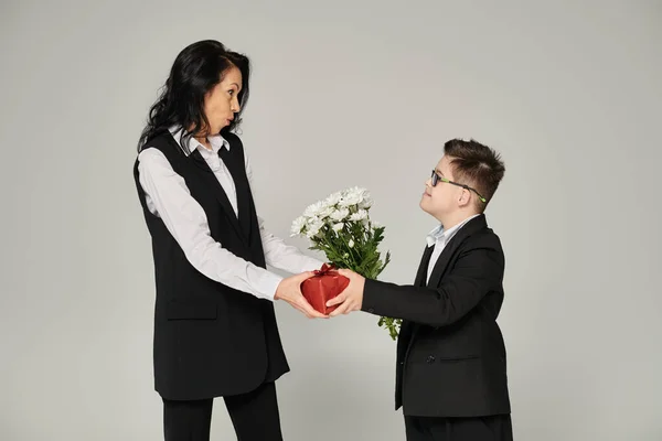 Schüler mit Down-Syndrom überreicht Blumen und Geschenkschachtel an überraschte Mutter auf grau — Stockfoto