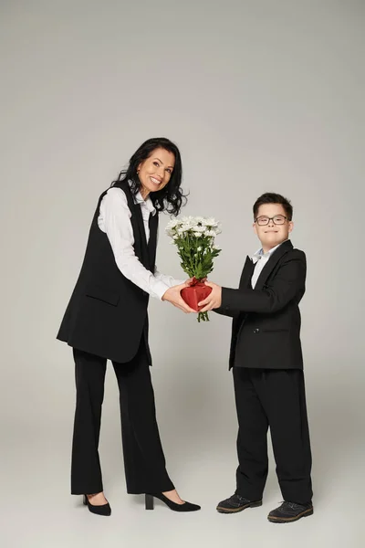Happy businesswoman and son with down syndrome holding gift box and flowers on grey — Stock Photo