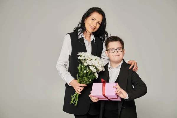 Elegant woman and boy with down syndrome with flowers an gift box smiling on grey, special family — Stock Photo