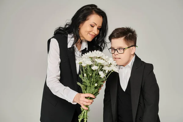Mulher alegre segurando flores perto do filho com síndrome de down em uniforme escolar e óculos em cinza — Fotografia de Stock