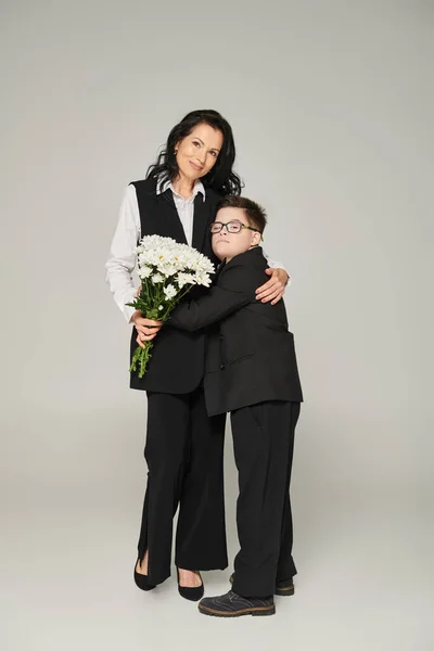 Happy woman and boy with down syndrome holding flowers and hugging on grey, unique family — Stock Photo