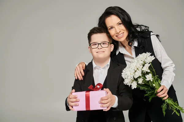 Felice donna e figlio con la sindrome di Down contenente fiori e scatola regalo, sorridente alla fotocamera su grigio — Foto stock