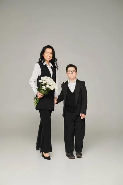 Woman in formal wear holding flowers near son with down syndrome in school uniform on grey — Stock Photo