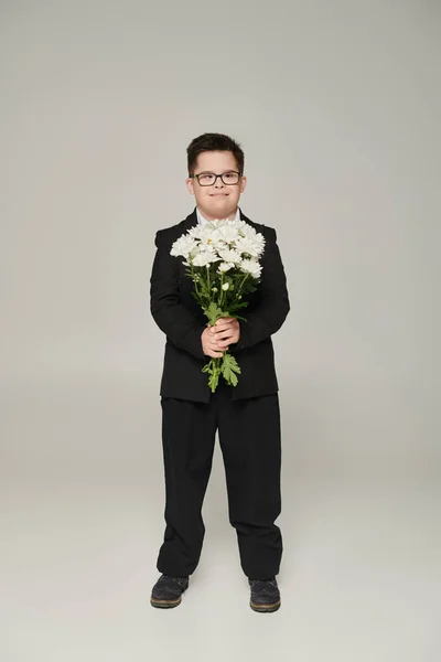 Garçon avec le syndrome du duvet en uniforme scolaire et lunettes tenant des fleurs et souriant sur gris — Photo de stock